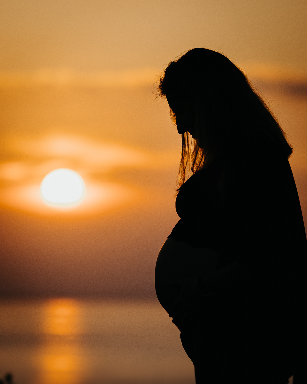 Silueta de mujer embarazada en contraluz, de pie frente a un fondo anaranjado de atardecer.

